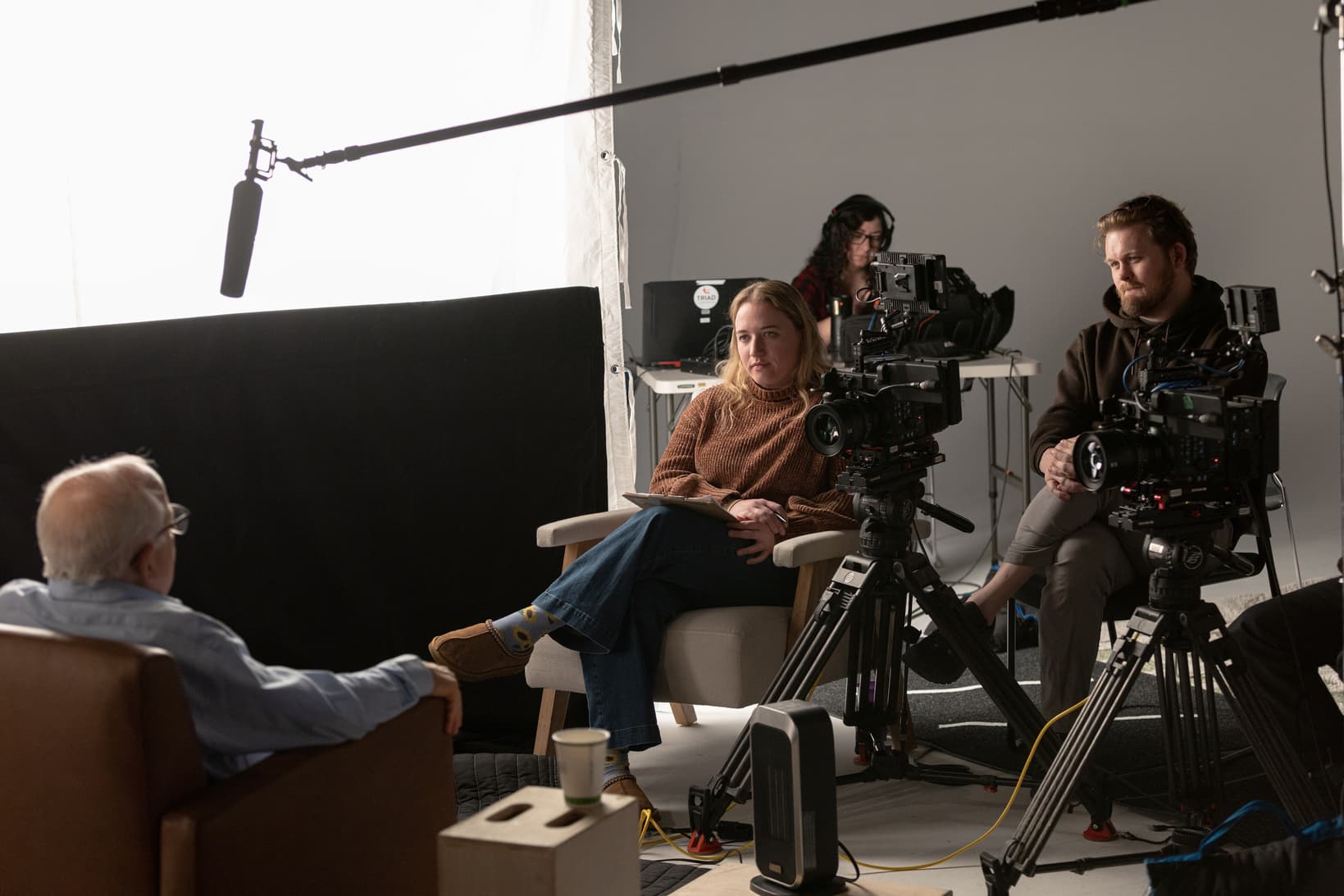 A film crew with two cameras and a boom microphone recording an interview with an older man in a studio