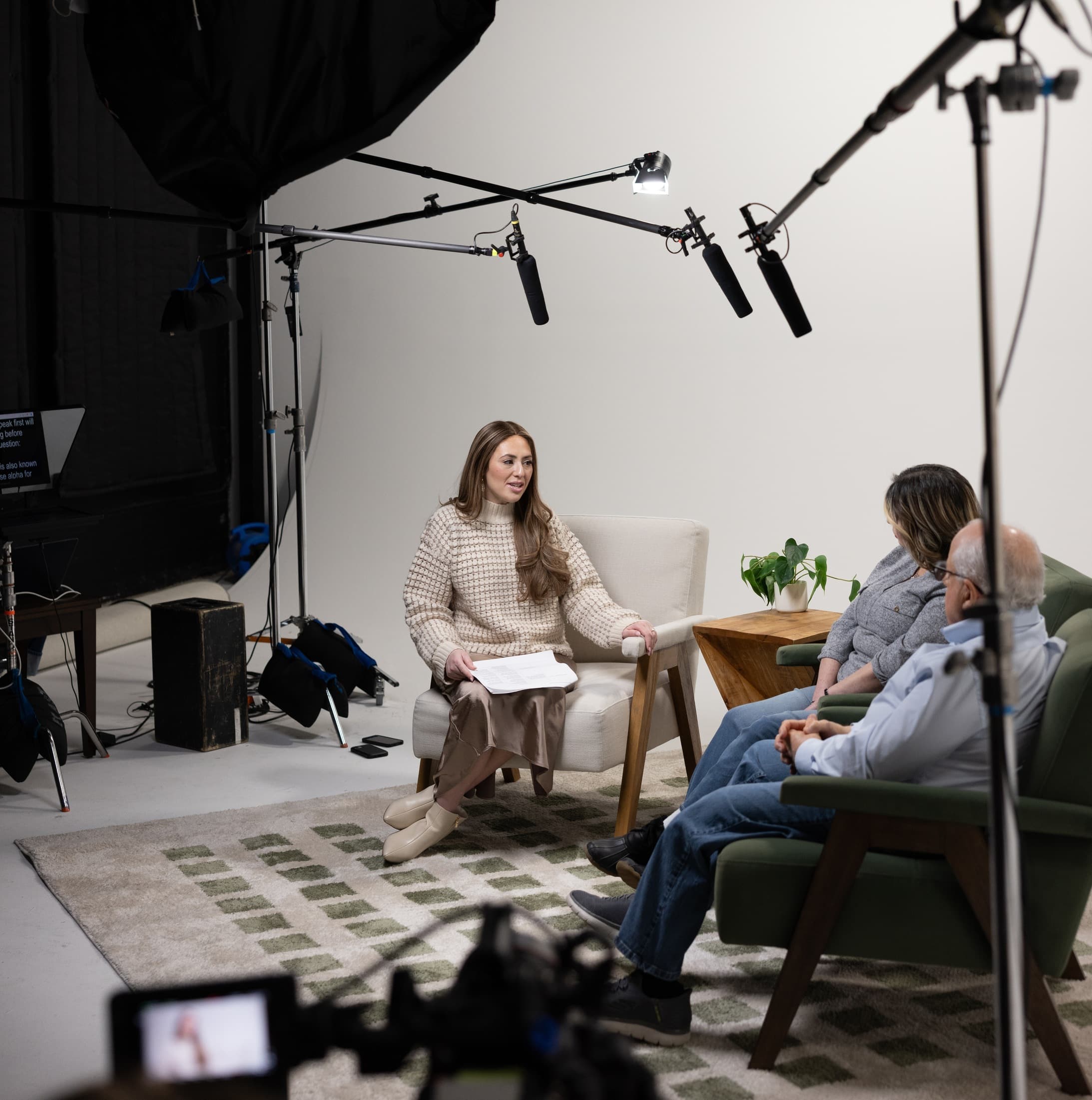 A presenter being filmed in a studio, with three boom microphones overhead and two participants listening